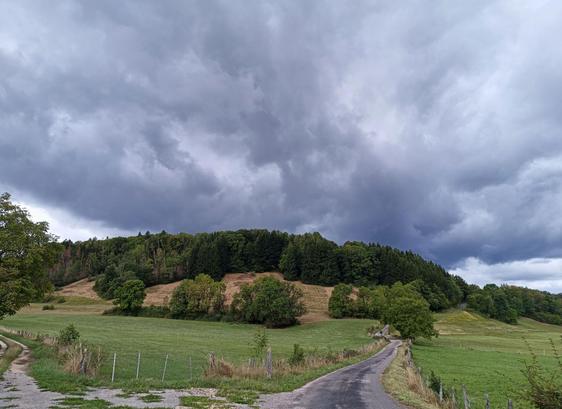 Photo. numérique couleur en format paysage, avec la vue large d'un paysage : au premier plan, l'intersection d'une route et d'un chemin, puis une prairie  des champs et des bois et au-dessus, un ciel chargé avec de gros nuages gris et bleu foncé en éventail.