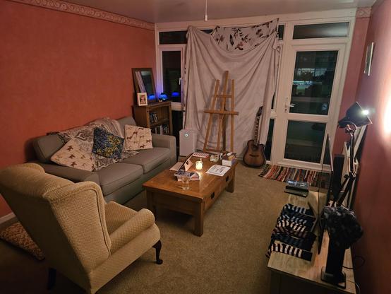 A photo of the living room. A mint green sofa faces the media centre, a grandma chair faces an easel and guitar. A bookcase filled with strange media is in the background, and a large coffee table is in the middle of it all. A large bedsheet is being used to cover the window because I am poor and curtains are expensive. Plus, I need to pick up the curtain rails from the delivery point.
