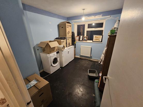 A photo of the spare bedroom. A naked floor (black molly tiles) are omnipresent, a washer, fridge, and freezer are alongside one wall with several boxes stacked on top. Frames of art are leaning against the window because I don't know how to hang a picture properly.