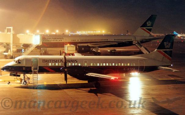 Night-time side view of a twin propellor-engined airliner parked facing to the left.
The plane has a dark blue lower fuselage, with the upper fuselage being dark grey, with black "British Airways" titles on the upper forward fuselage.
The grey registration "G-BTPD" is on the lower rear fuselage
The forward door is open, with a built-in set of airstairs deployed.
A larger, twin engined jet airliner in a similar livery is parked on the next gate, with an air bridge attached to the forward door.
Bright lights from airport lighting stands, vehicles, and other planes illuminate the scene, blowing out some parts of the image with the long exposure required to bring out details.