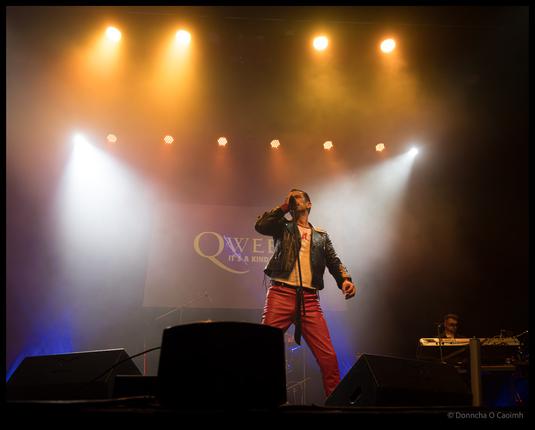 A Freddie Mercury tribute performer in red trousers, black leather jacket, and yellow shirt stands centre stage holding a microphone with one arm outstretched under dramatic white spotlights and golden stage lighting, with "QWEEN IT'S A KIND" visible on a backdrop at Cork Opera House.