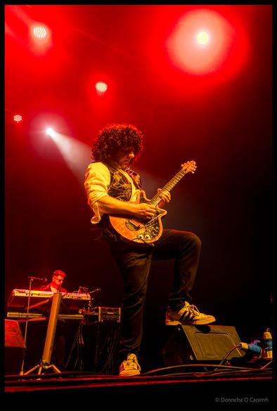 A guitarist with curly dark hair ala Brian May wearing a white shirt made yellow under the red lights and ornate waistcoat playing an electric guitar, with a keyboardist visible in the background at the Cork Opera House during a Qween tribute band performance
