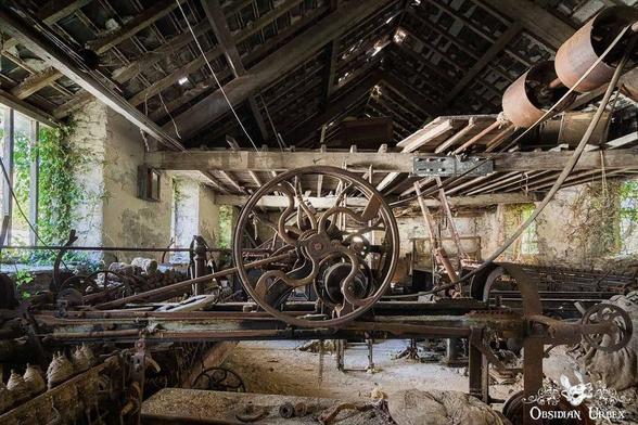 Old, rusted machinery with large gears and belts inside an abandoned, dilapidated building with overgrown plants visible through broken windows.