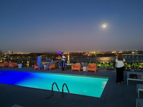 Picture of a hotel rooftop swimming pool, illuminated blue and green, against an urban skyline backdrop, and a river shimmering by the light of a full moon in a cloudless sky.