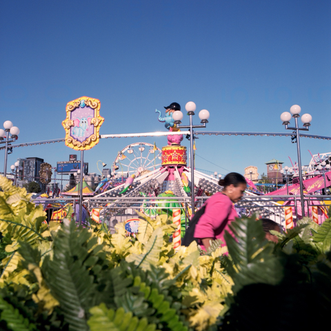 A woman walks past a cluster of plastic ferns with the kids’ midway behind her.