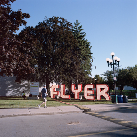 A quiet moment as a man walks pasts an old fashioned illuminated sign that spells “FLYER”, those of a certain age will recognize the sign from the somewhat infamous wooden roller coaster from The Ex of yore. A security guard sits in the shade nearby.