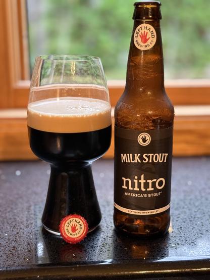 A stout glass full of dark beer with a thick tan head. The red and white bottle cap with a red left hand is balanced in front, and the brown bottle with a black and white label is to the right