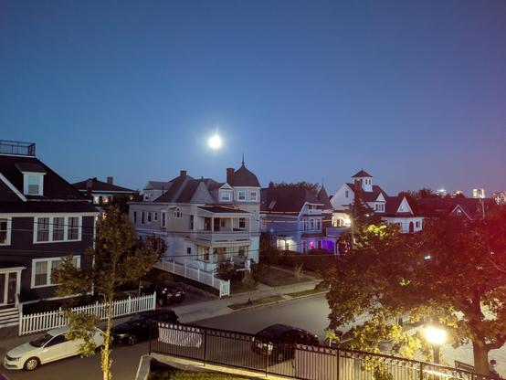 the moon in the sky over houses on top of a hill. you can kinda see Boston off to the right