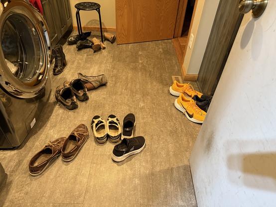 9 pairs of shoes are strewn across a gray floor just inside an opened door. a front loader washing machine is to the left