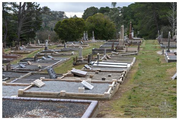 An overhead wide angle shot of a cemetery showing a gentle slope covered in numerous graves and headstones.

The closest graves are concrete surrounds filled with grey gravel, with several flat or slightly tilted memorial plaques placed within them.

The rows of graves extend back up the hill, with various styles of monuments and weathered upright headstones visible. 

The far background features a dense line of lush green trees on the hillside under a bright, cloudy sky.