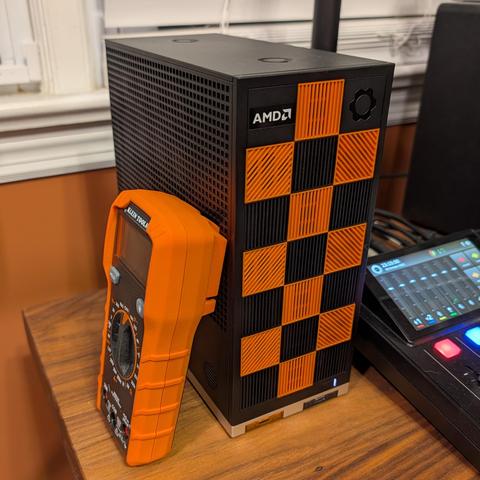 A Framework Desktop computer with black and orange front tiles arranged in a checkered pattern. There's an orange handheld multimeter leaning on the desktop showing how small the desktop is.