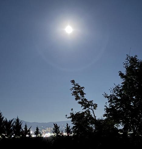 A bright full moon is visible in a clear night sky, surrounded by a soft halo. Silhouetted trees frame the scene, with distant city lights glimmering below the horizon.
