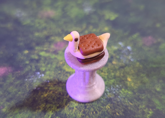 tiny figurine of a white duck that's been squished between two graham crackers with chocolate, like a marshmallow in a s'more. it's sitting on top of a tiny stone pedestal, against a grassy background.