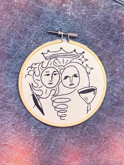 Un aro redondo bordado con dos caras abstractas y una corona encima, entrelazadas con una forma en espiral, simbolizando al soy y la luna. El fondo es de mezclilla texturizada, lo que añade profundidad a la imagen.