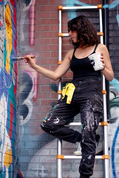 A female street artist standing elegantly on a ladder, painting a wall with a brush.