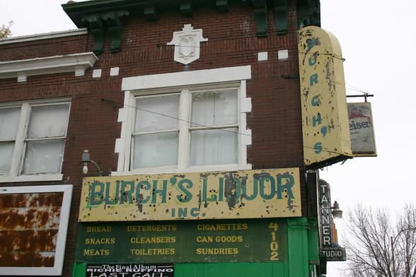 Photo of the exterior of a corner store. A green on yellow sign on the wall facing the camera reads "BURCH'S LIQUOR INC." in ornamented capitals. Projecting from the corner of the building is another green on yellow sign with a half-circle top that reads "BURCH'S" in sans-serif capitals.