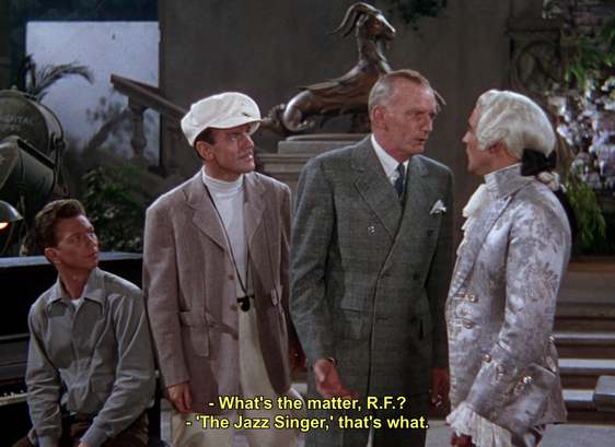 A group of men stand around a film stage, one in an elaborate costume. One says, "What's the matter, R.F.?" He replies, "The Jazz Singer, that's what."