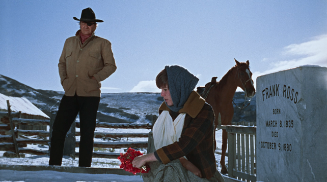 Amidst a snowy landscape, a cowboy with an eyepatch stands over a young girl, who is placing flowers on a grave. The gravestone reads "Frank Ross, born March 8, 1835, died October 6, 1880"