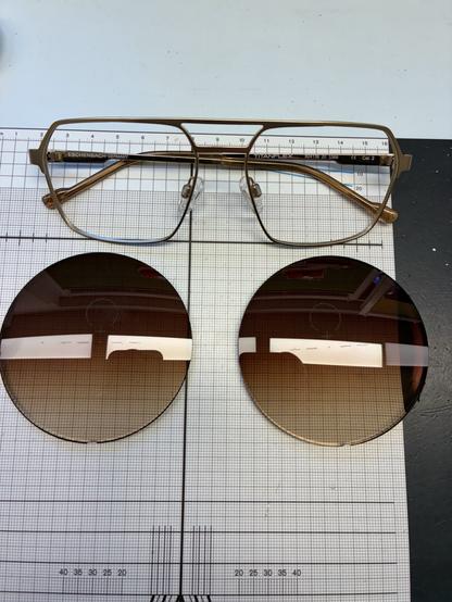 Eine goldene Doppelsteg-Brillenfassung auf einer Arbeitsunterlage mit Millimeter-Raster. Da drunter liegen die noch ungeschliffenen Rohgläser mit Braun-Verlauf