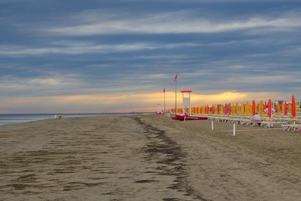 A wide, peaceful beach stretches into the distance under a cloudy blue-grey sky. The sea lies calm on the left, while the right side of the image is lined with neat rows of closed beach umbrellas in bright orange, yellow, and red. A red lifeguard tower and rescue boat stand out among the sun beds, their colours vivid against the muted tones of the sand and sky. Two small figures walk along the shoreline far in the distance. The horizon glows softly with the warm light of sunset, adding a gentle golden hue to the tranquil scene.