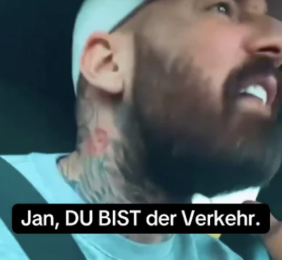 Screenshot aus dem Video, das verlinkt ist. Du sienst einen tätowierten Mann mit türkiser Kappe und Tshirt angeschnallt im Auto angestrengt gucken. Darüber steht: Jan, du BIST der Verkerh.