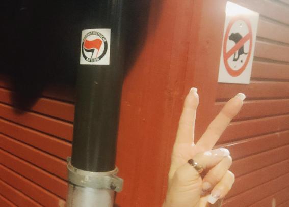 Hand with long nails and thumb ring making V for victory in front of 2 black-red-white signs on wooden building 
- black dog defecating, very graphic, with red ring and slash, no dog shit
- sticker with Antifa logo (2 sign-less flags) and text in a ring around it: 
ANTIFASCIST ACTION 

Photo, sticker and hand by me