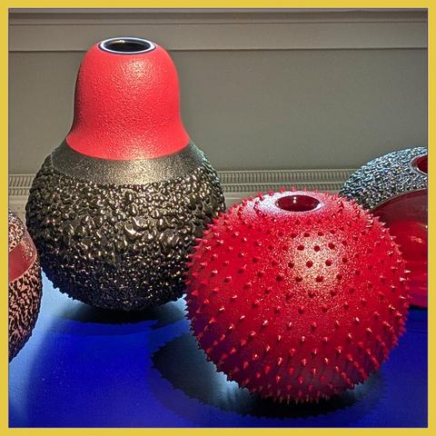 Two colorful round vases at a dark blue table. First vase is red, with little pins on it. Second vase is red and dark gray