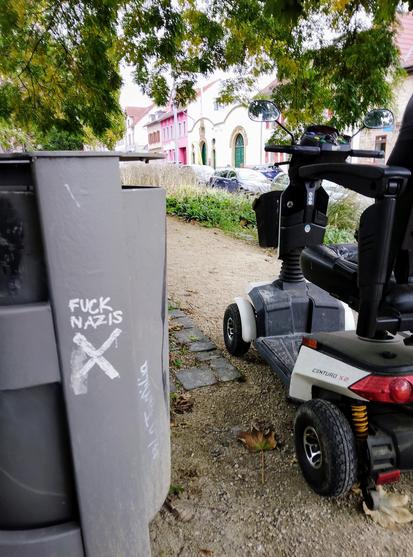 The scoot-chair is parked next to a trash can emblazoned with FUCK NAZIS, and the Shitter logo, which is a sans-serif swastika.