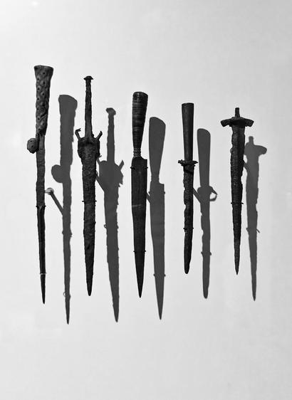 A photo of row of ancient black metal daggers displayed hanging upright in front of a bright background, each casting a distinct shadow on the wall behind.