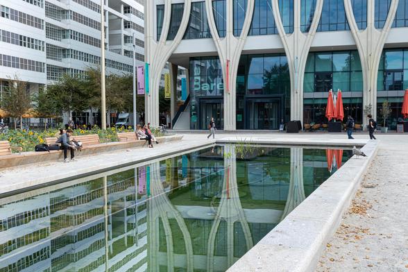 Photo of a modern, rectangular pond with a white stone edge, taken lengthwise, diagonally from left to right. Around it are wooden benches, on which a few people are seated. Behind the pond, the lower part of the facade of The Hague Concert Hall can be seen, with four pillars that fan out, interspersed with glass windows. On the left is the white City Hall with rows of windows. The buildings are reflected in the water of the pond.