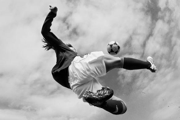 A female soccer player in sportswear performs a bicycle kick. She is wearing a long-sleeved jersey, shorts, and long socks. The camera angle is from the ground looking up, with the cloudy sky visible in the background. The soccer ball is hovering near her outstretched leg.