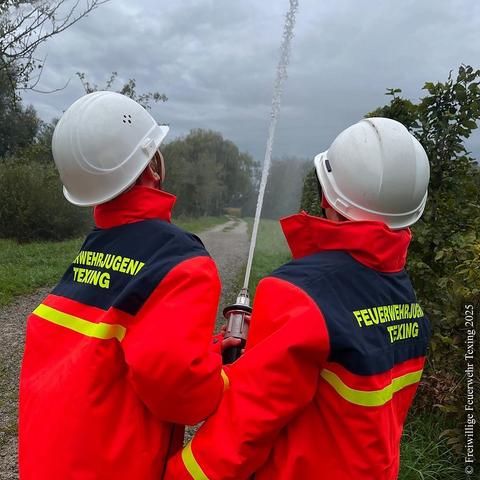 Zwei Mitglieder der Feuerwehrjugend Texing-St. Gotthard von hinten. Die beiden Buben tragen jeweils einen weißen Schutzhelm und rote Schutzjacken der Feuerwehrjugend Texing. 
Sie halten gemeinsam ein Strahlrohr und spritzen Wasser in die Luft.

English: 
Two members of the Texing‑St. Gotthard youth fire brigade seen from behind. Each boy wears a white protective helmet and a red youth‑firefighter jacket. Together they hold a water‑stream nozzle and spray water into the air.