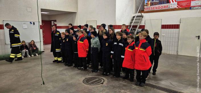 Die Mitglieder der Feuerwehrjugend Diesendorf und der Feuerwehrjugend Texing-St. Gotthard sind gemeinsam im Feuerwehrhaus der freiwilligen Feuerwehr Diesendorf zum Beginn der Veranstaltung angetreten. Im Hintergrund sieht man zwei Jugendbetreuer, zwei Kinder ohne Uniform sitzen auf Sandsäcken und beobachten das Geschehen.

English: 
The members of the Diesendorf youth fire brigade and the Texing‑St. Gotthard youth fire brigade have gathered together in the volunteer fire station of Diesendorf at the start of the event. In the background two youth supervisors and two children without uniforms are sitting on sandbags, watching the scene.