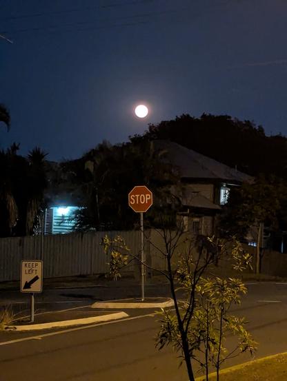 A full moon rises above trees and a house, with a STOP sign in the foreground.