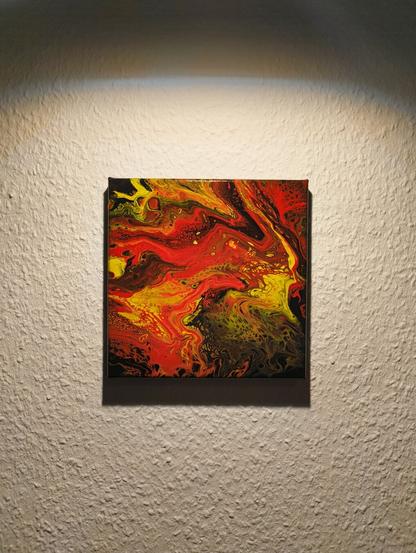 Das Foto zeigt ein abstraktes, quadratisches Acryl-Gemälde mit intensiven Rot-, Gelb- und Schwarztönen, das an einer hellen, strukturierten Wand hängt und von oben angeleuchtet wird. Das Gemälde besteht aus abstrakten Farbverläufen und Mustern, die an fließende Lava oder bewegte Flüssigkeit erinnern. Die dominierenden Farben sind Rot, Gelb, Orange und Schwarz, mit leichten Übergängen ins Grünliche. Die Oberfläche ist glänzend lackiert.