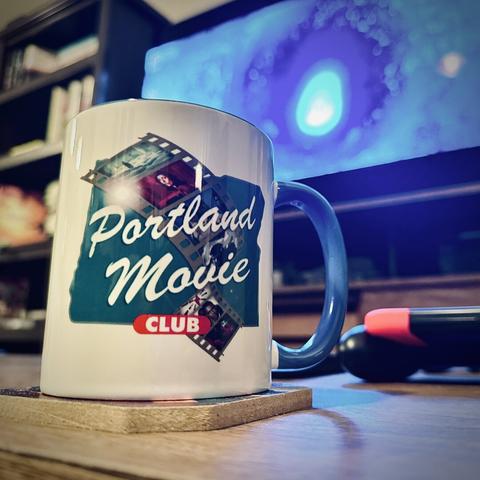 A white and blue coffee mug with the Portland Movie Club logo sits on a coffee table in the foreground, as a large television next to a bookcase in the distance plays a bright blue scene from a science fiction film.