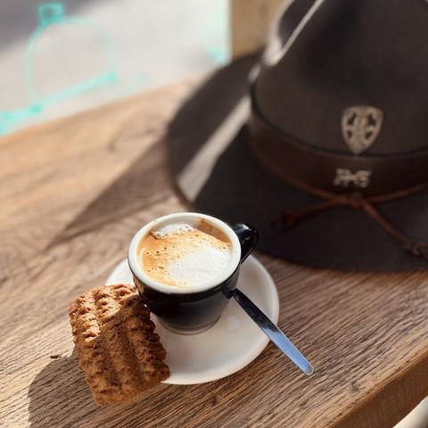 Still time for another coffee at SlowWings, in Ypres.
A cup of coffee on a wooden bar, a scout hat in the background.