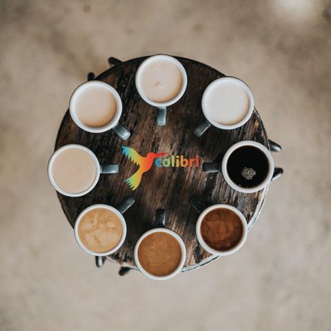 Logo Café Colibri in der Mitte. Auf einem runden Holzbrett stehen Kaffeetassen mit verschiedenfarbigem Inhalt.