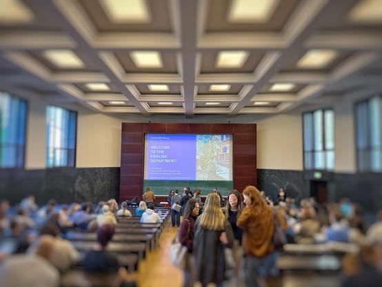 A large lecture hall. In the distance, a screen with a welcome message. Students are seated along rows of desks, with a few still standing in the aisles.