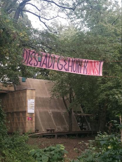 Wir sind am Osthang.
Im Hintegrund sieht man die Mainhall: Ein organischa ussehendes gebäude aus holz. Umringt ist es von Bäumen.
von dem gebäude, durch die bäume geht ein weg zu uns.Über dem Weg hängt ein Rosa Banner auf dem in lila Schrift steht "Die Stadt Gehört Kunst", wobei das "uns" in "Kunst" in einem pink ist. und Somit als "(K)uns(t)" gelesenw erden kann.