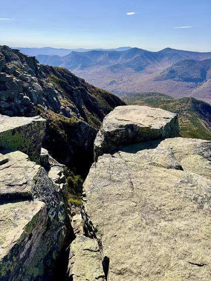Photo of flat slabs of rock at edge of cliff while looking at distant mountain range. The slabs are split in two pieces with crack between the two showing view of valley. Blue sky overhead.

Bondcliff Mountain with view of Franconia Ridge.