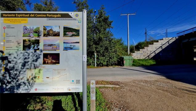 En stor skylt omgärdad av lövträd. I bakgrunden en väg. På höger sida fortsätter vägen under en betongbro.