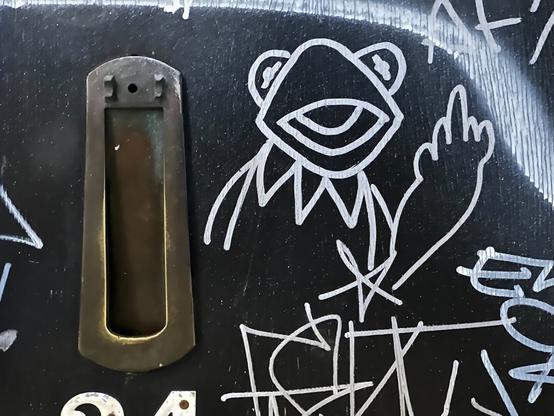white paint marker graffiti on a black door. Amongst the scrawled tags is a drawing of the Muppet character Kermit The Frog giving the middle finger