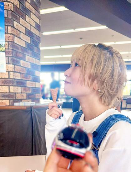 A woman with short blonde hair sits indoors, holding an ice cream cone and looking thoughtfully toward a brick pillar. She is dressed casually in a white shirt and denim overalls. In the foreground, a hand holds a small black and red round object, slightly out of focus.