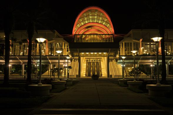 During the night, the Orlando convention Center shines neon orange lights. Rounded street lamps light the scene in the foreground.