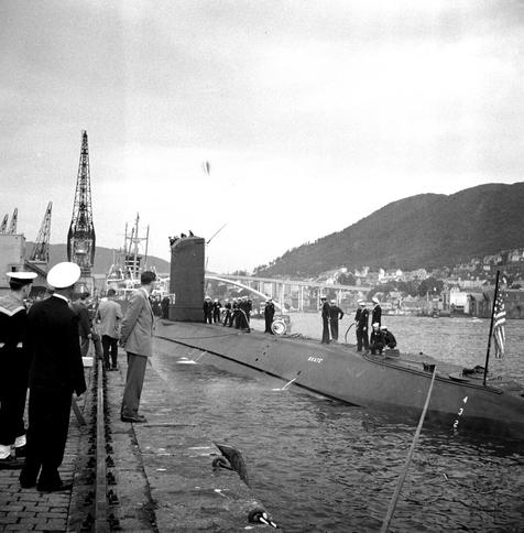 A black and white image of an American submarine docked in a Bergen fjord. In the background, mountains and the Puddefjord bridge.