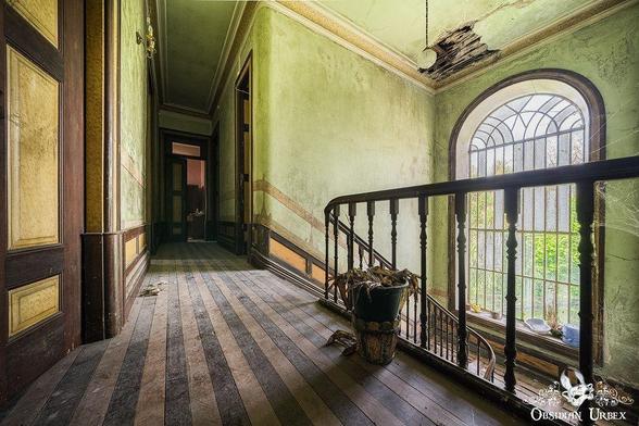 An abandoned upper hallway with peeling green walls, damaged ceiling, wooden railing, potted plant, and large arched window letting in natural light.