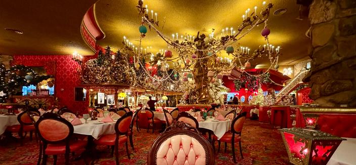 A large dining room that has Christmas decorations galore. There is a pole in the center of the room that serves as a regular type tree, and it is covered with lights, ornaments and candles. Across the room is an upper ledge near the ceiling that has several Christmas trees full of lights and decorations. The decor of the room is red and pink (that is their signature look). The chairs are pink vinyl cushions and the floor is dark pink with a flower motif. The walls are red. There is just too much to describe!   Madonna Inn, San Luis Obispo, California.