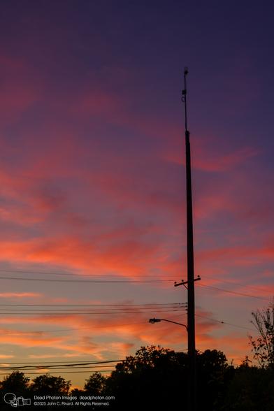 A photograph of a tall utility pole silhouetted against a vibrant sunset sky with hues of orange, pink, and purple. Trees and power lines are silhouetted at the bottom.