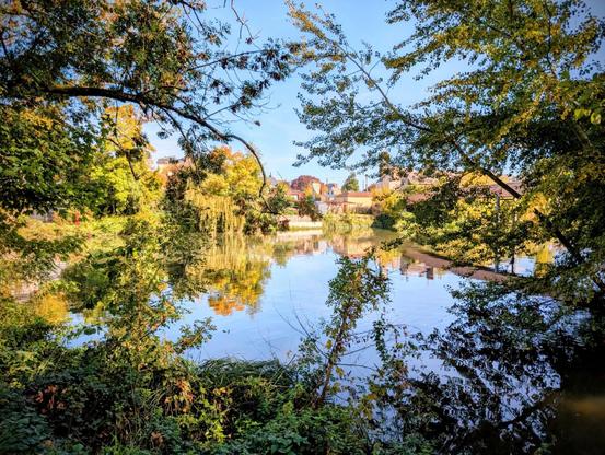 Vue sur la rivière depuis le parc avec une pièce d'eau entourée d'arbres et de feuillages avec un beau ciel bleu qui se reflète.